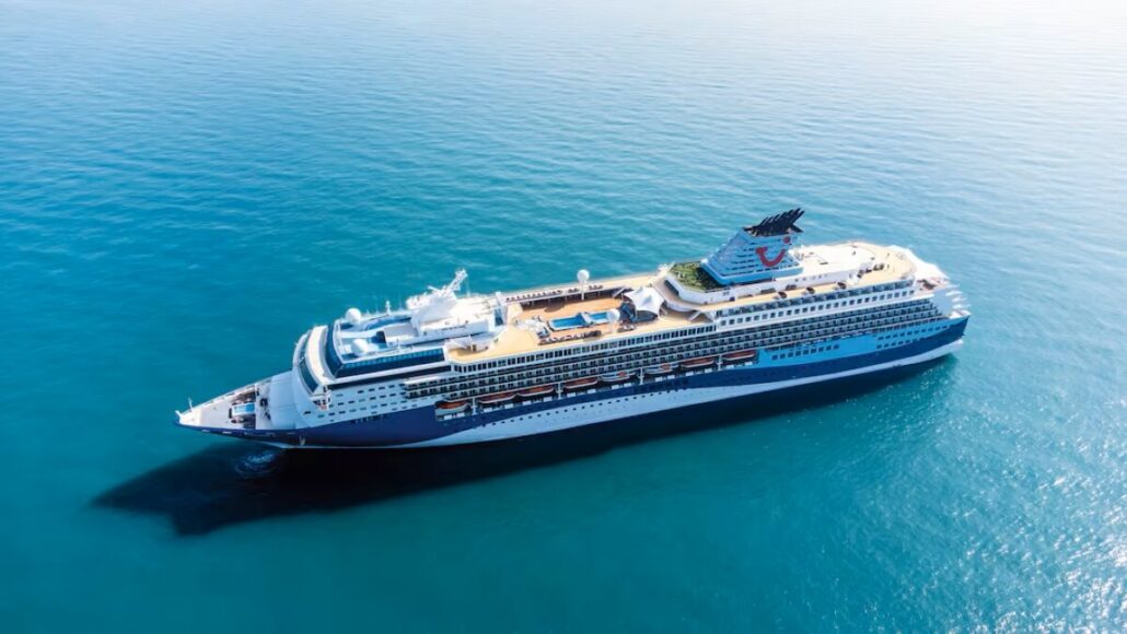 A large white and blue cruise ship sails on calm, clear blue water under a bright sky. The upper decks feature outdoor seating areas, a pool, inviting staterooms for guests, and lifeboats visible on the sides.