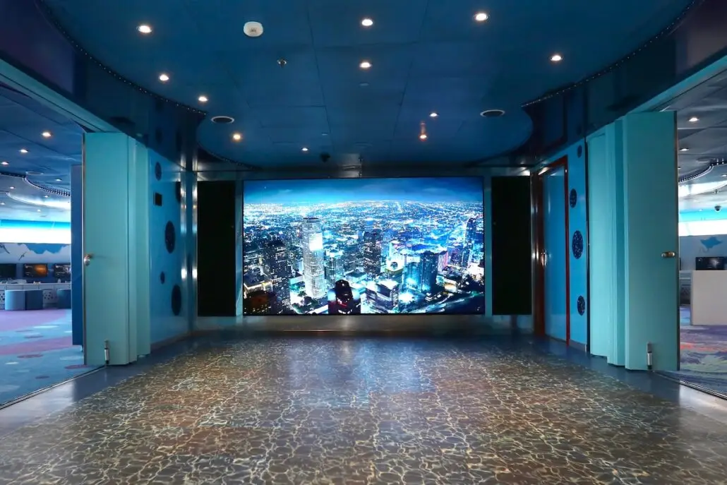 A hallway with blue walls and a patterned floor leads to the Tribute Lounge, where a large screen displays a nighttime cityscape with tall buildings and bright lights. The ceiling is also blue with recessed lighting, evoking the vibrant ambiance of Carnival Valor.
