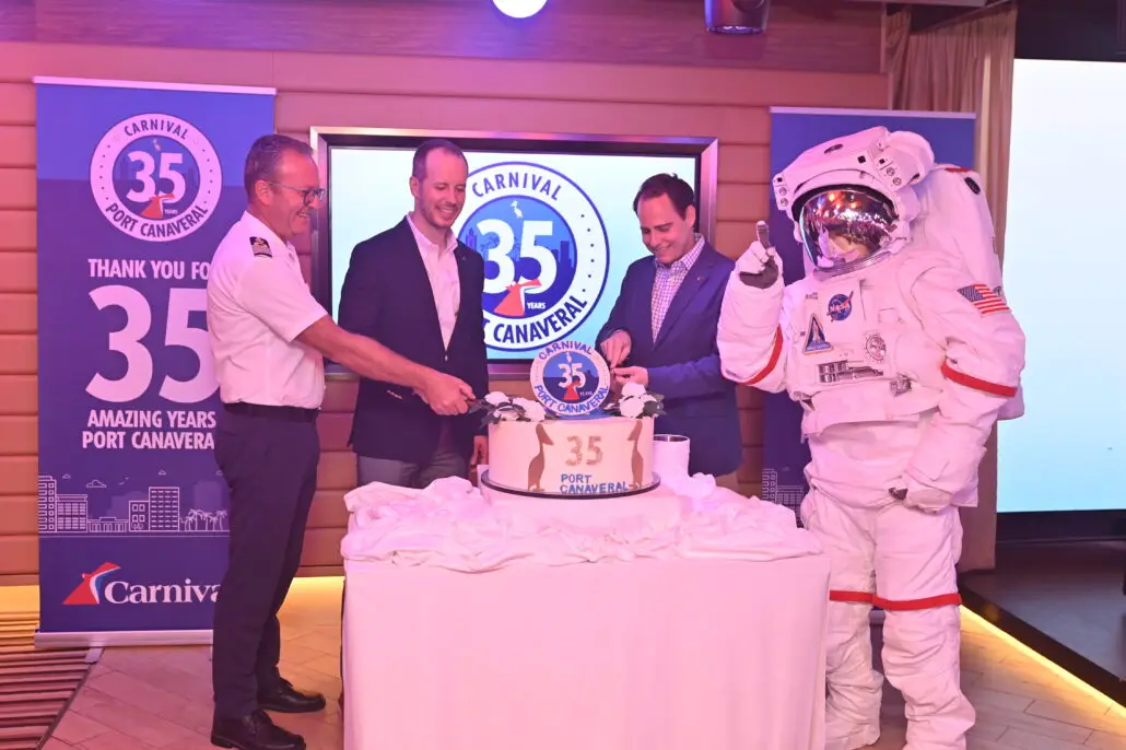 Four people—three in business attire and one in an astronaut suit—stand around a cake marked “35 Port Canaveral,” celebrating Carnival’s 35 Years at Port Canaveral; two banners decorate the background.
