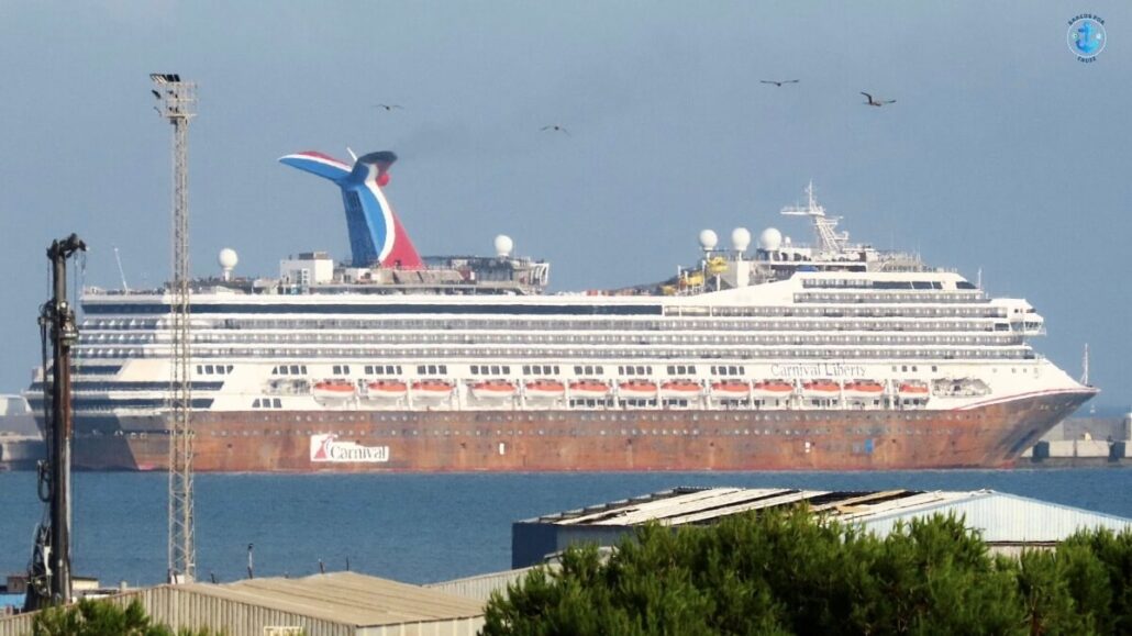 A large white Carnival cruise ship with multiple decks, lifeboats, and a red, white, and blue funnel is docked at a port. The weathered hull hints at a recent drydock delay in Spain Shipyard, as birds fly in the sky above.