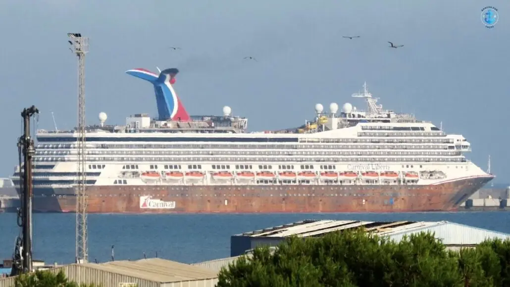 A large white Carnival cruise ship with multiple decks, lifeboats, and a red, white, and blue funnel is docked at a port. The weathered hull hints at a recent drydock delay in Spain Shipyard, as birds fly in the sky above.