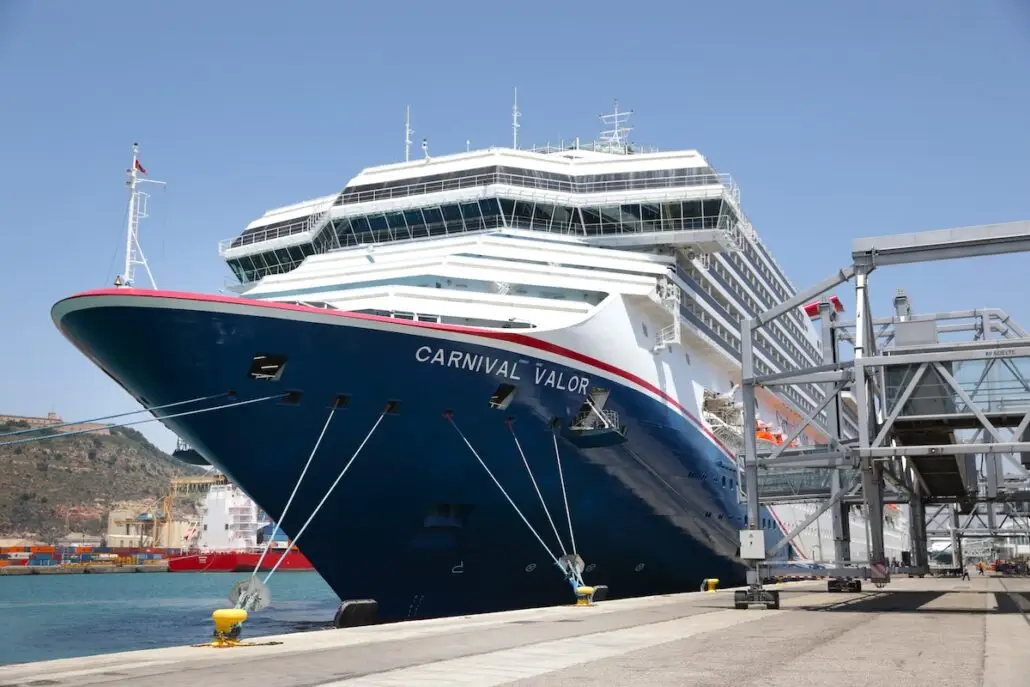 A large cruise ship named Carnival Valor, featuring attractions like the Tribute Lounge, is docked at a port with a boarding bridge attached and industrial structures visible in the background under a clear sky.