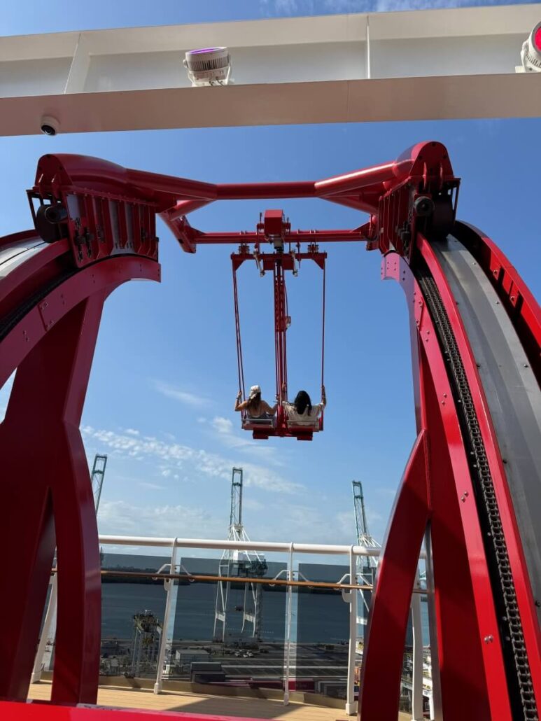 Two people enjoy a red swing ride suspended in the air on MSC World America’s deck, with blue sky, cranes, and water in the background—perfect for unforgettable moments on a family cruise.