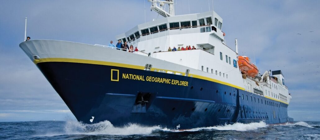 A large blue and white ship labeled "National Geographic Explorer" sails on the ocean under a cloudy sky during an Antarctica Expedition Cruise, with people standing on deck and orange lifeboats visible on the side.