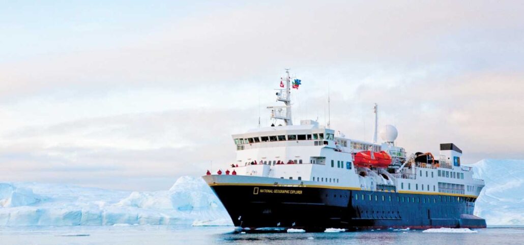 A large Antarctica Expedition Cruise ship with a blue hull and white superstructure sails through icy waters, surrounded by snow-covered icebergs and cloudy skies in the background.