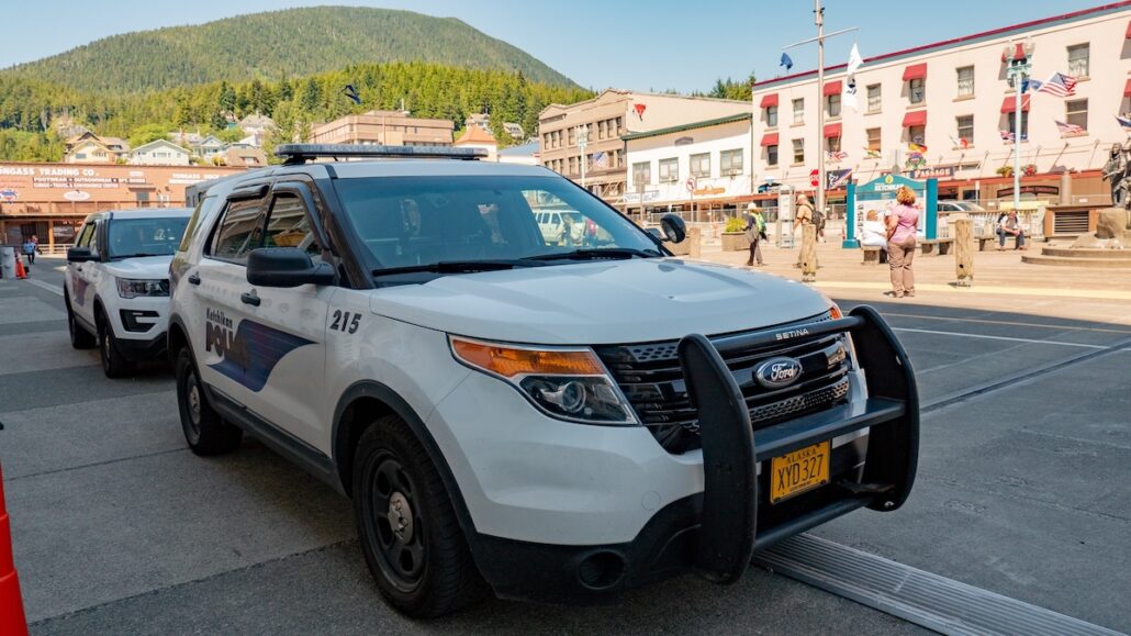 A white police SUV with a push bumper is parked on an Alaska street in front of buildings. Another police vehicle is behind it, while people walk nearby and a forested hill rises in the background.