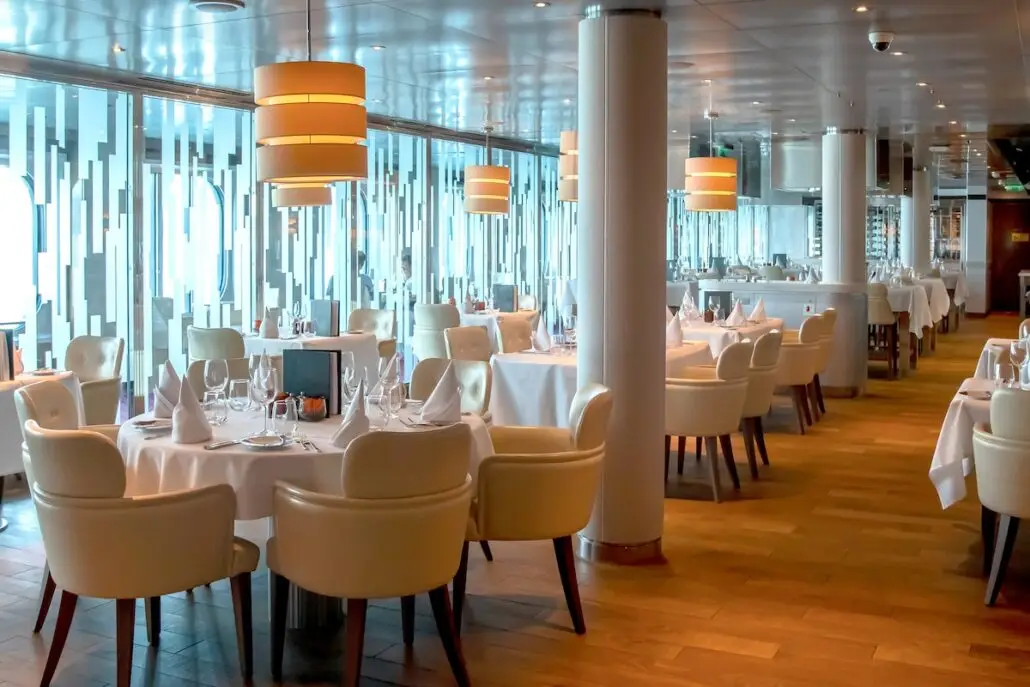 A modern restaurant aboard Koningsdam features white tablecloth-covered tables, cream-colored padded chairs, wooden floors, large windows, hanging pendant lights, and glass partitions with vertical frosted patterns.