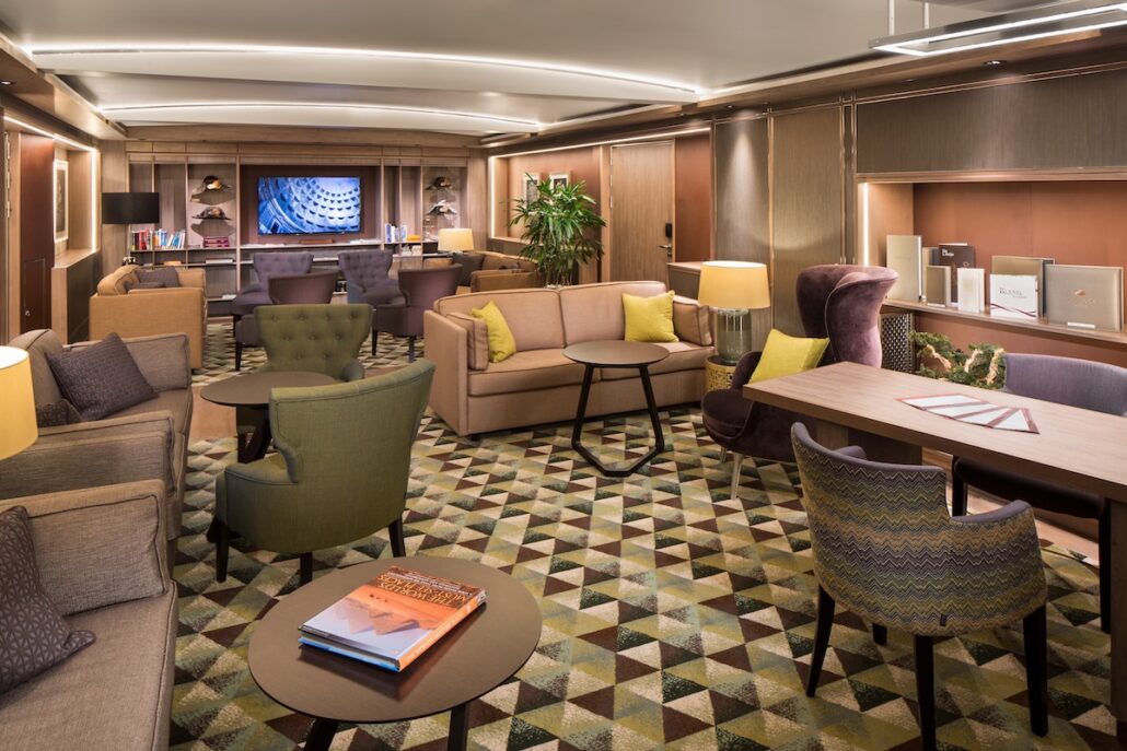 A modern lounge aboard Holland America’s Koningsdam, featuring patterned carpet, various armchairs, sofas and tables, soft lighting, bookshelves, decorative plants, and a television mounted on the far wall—perfect for Neptune Suite guests.