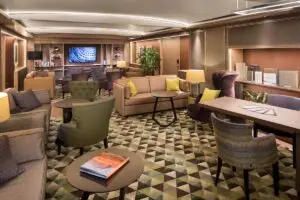 A modern lounge aboard Holland America’s Koningsdam, featuring patterned carpet, various armchairs, sofas and tables, soft lighting, bookshelves, decorative plants, and a television mounted on the far wall—perfect for Neptune Suite guests.