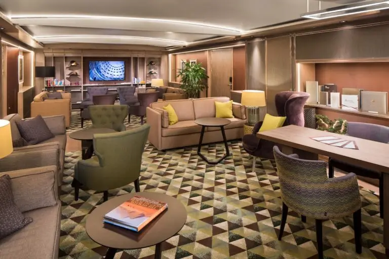 A modern lounge aboard Holland America’s Koningsdam, featuring patterned carpet, various armchairs, sofas and tables, soft lighting, bookshelves, decorative plants, and a television mounted on the far wall—perfect for Neptune Suite guests.