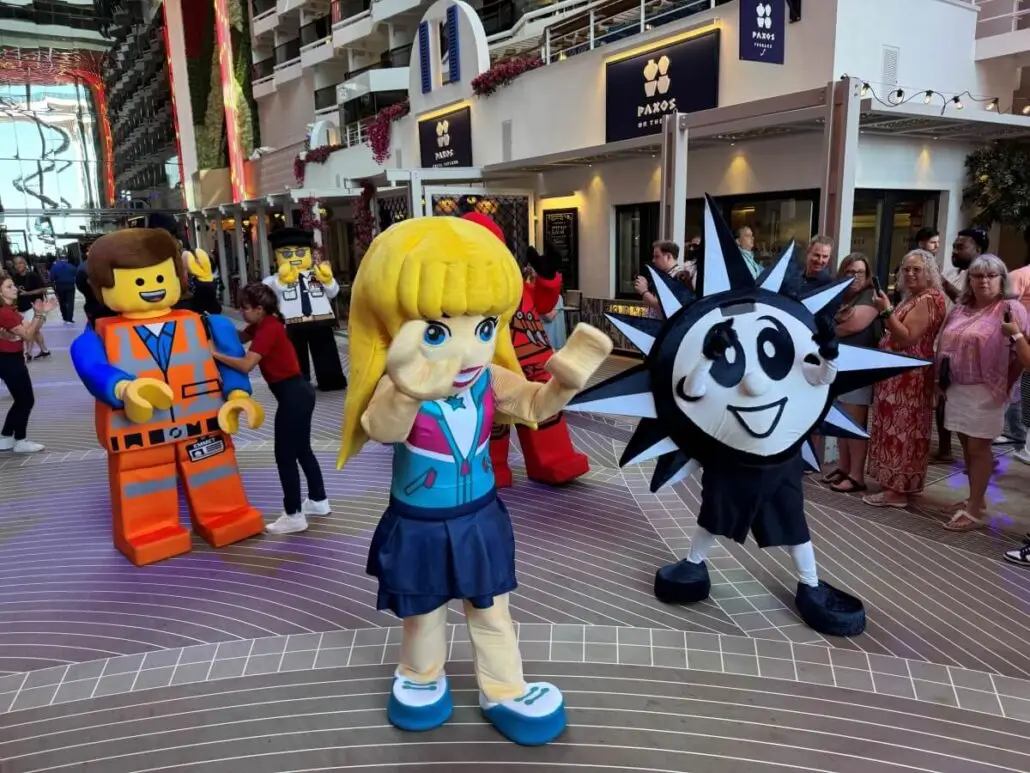 People in large costumes, including characters resembling LEGO figures and a black-and-white sun mascot, entertain a crowd of onlookers inside a spacious indoor area with balconies and shops—perfect for families cruising on MSC World America.