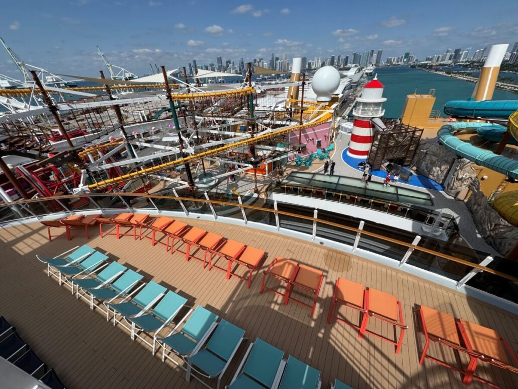 Rows of orange and turquoise lounge chairs on MSC World America’s deck overlook a ropes course, a red and white lighthouse structure, water slides, and a city skyline—perfect for a family cruise under a partly cloudy sky.