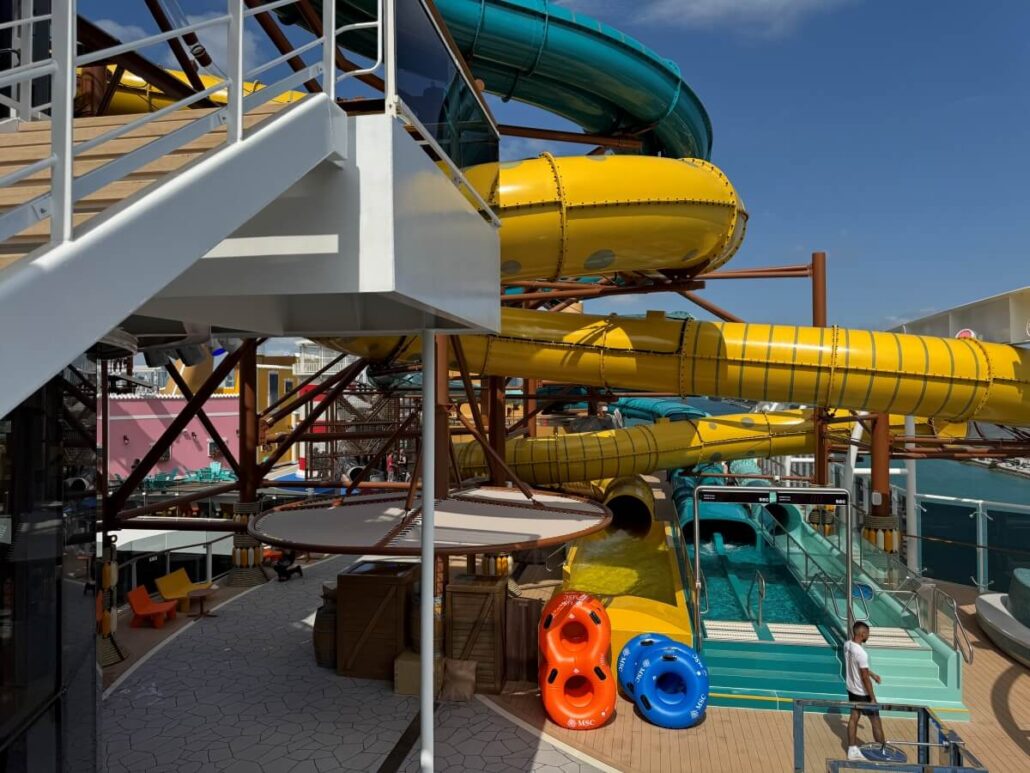 A bright outdoor water park aboard MSC World America features yellow and teal tube slides, a staircase, lounge chairs, two inflatable rafts, and a person near the slide exit—perfect for discovering what's new on MSC ships under a clear blue sky.
