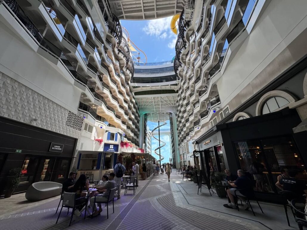 People sit at tables in an open-air atrium lined with shops and balconies on both sides—a highlight among new MSC ship features. Sunlight pours through the partially covered roof, with a spiral slide at the far end under a blue sky.