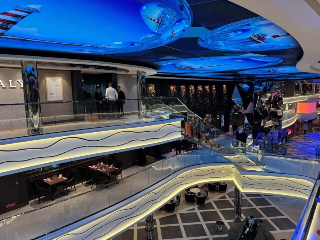 Interior view of a modern multi-level shopping mall aboard MSC World America, featuring wavy-lit railings, glass balconies, dining areas on each floor, and a striking blue digital ceiling display—showcasing signature cruise ship features.