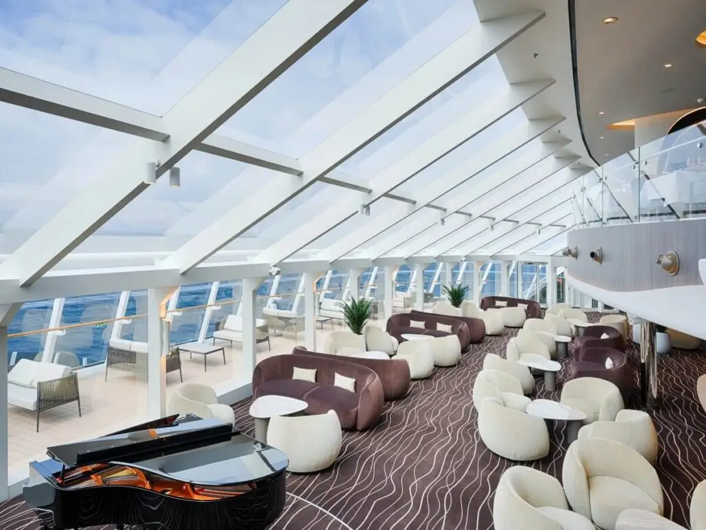 A modern lounge area on MSC World America features floor-to-ceiling windows, curved sofas, white chairs, small tables, a grand piano, and stunning ocean views—showcasing the new features found on the latest MSC ships.