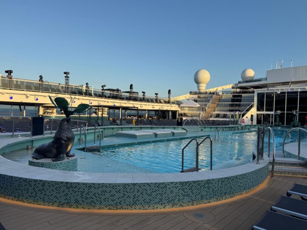 An outdoor pool on MSC World America’s deck features a whale tail sculpture at one end. Surrounded by lounge chairs and glass railings, this area showcases the new features found on MSC ships, with radar domes and upper decks visible in the background.