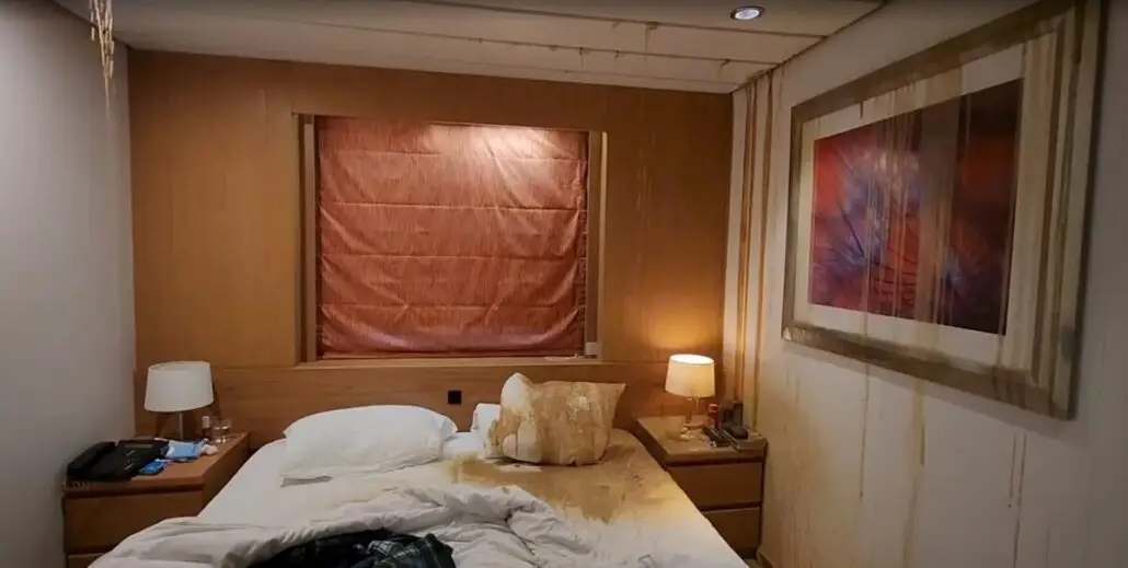 A small stateroom bedroom with a bed and two nightstands. The unmade bed has pillows askew and stained linens. Water stains mark the walls and ceiling, and even the artwork bears damage—far from a dream cruise vacation retreat.