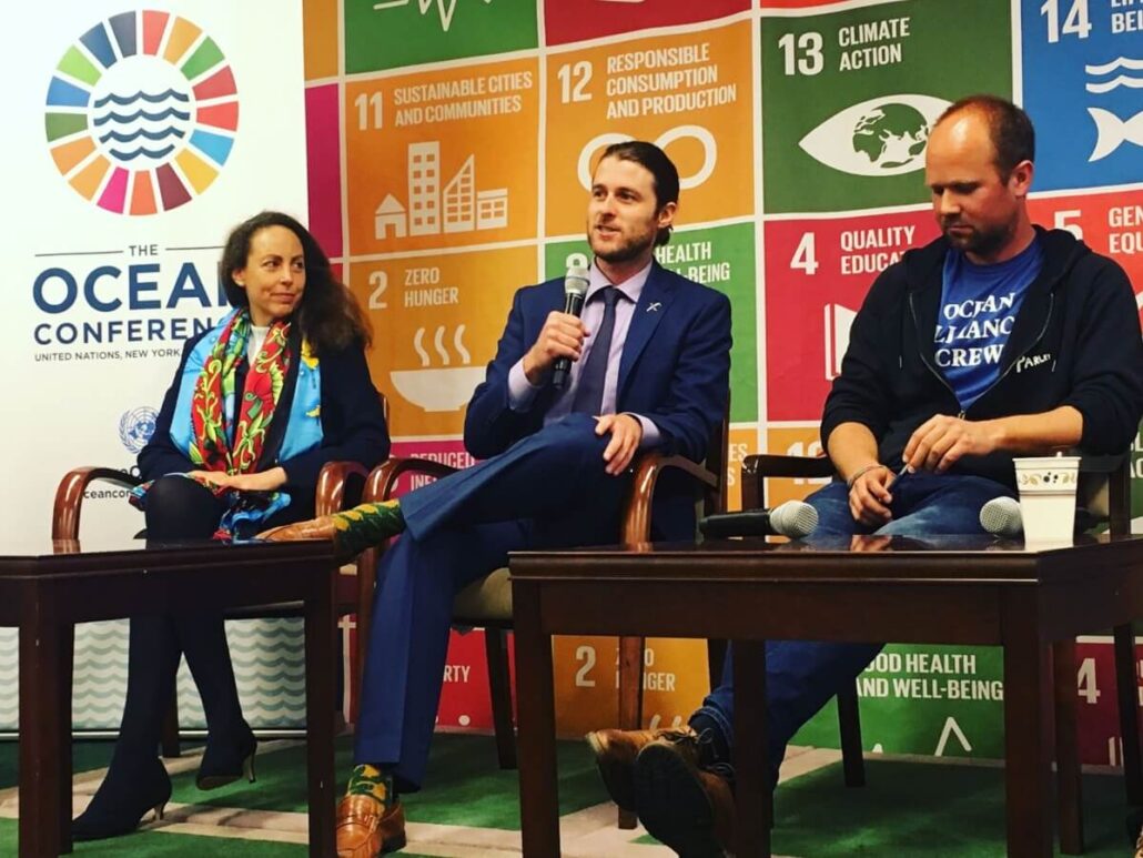 Three people sit on a stage in front of colorful UN Sustainable Development Goals icons at The Ocean Conference. The man in the center holds a microphone to discuss the environmental impact of Antarctica cruise ship tours while the others listen.