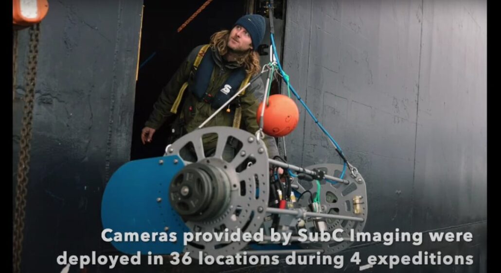 A person with long hair and a beanie monitors a large underwater video camera rig with mechanical parts and orange floats, being lowered over the side of a vessel. Text reads: "Cameras provided by SubC Imaging were deployed in 36 locations during 4 expeditions.