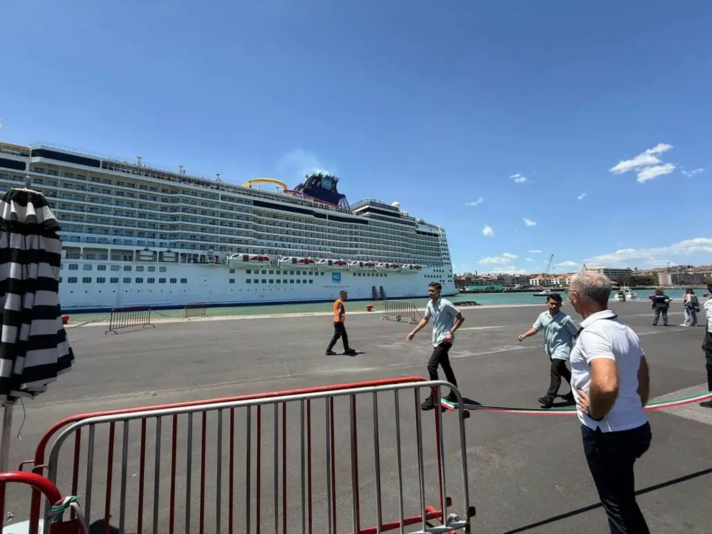A large cruise ship docked at a port; several people stand or walk on the pavement in front, with red metal barriers and a black-and-white striped umbrella in the foreground after strong winds swept through under a clear blue sky.