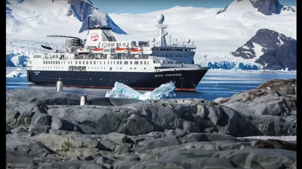 A large Antarctica cruise ship sails through icy waters near Antarctica, surrounded by snow-covered mountains and glaciers. Rocky terrain with a few penguins and ice chunks is visible in the foreground.