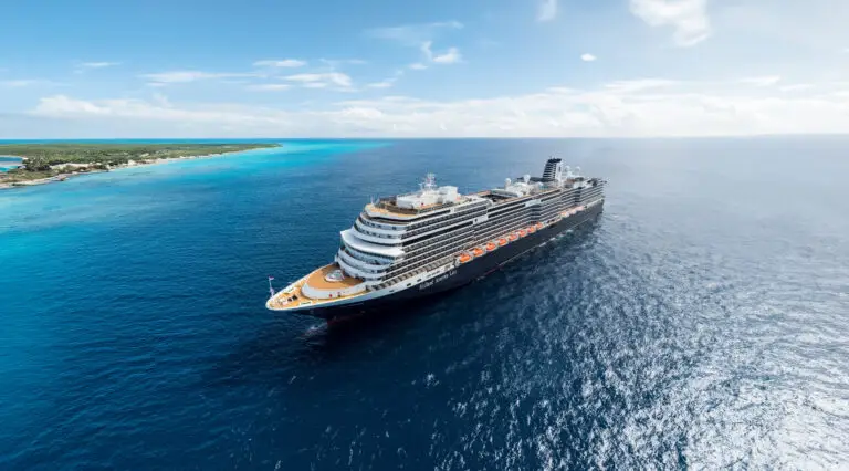 A large Holland America cruise ship sails on calm blue ocean water near a tropical coastline with clear skies and scattered clouds overhead, offering unforgettable cruises—don’t miss the latest sale.