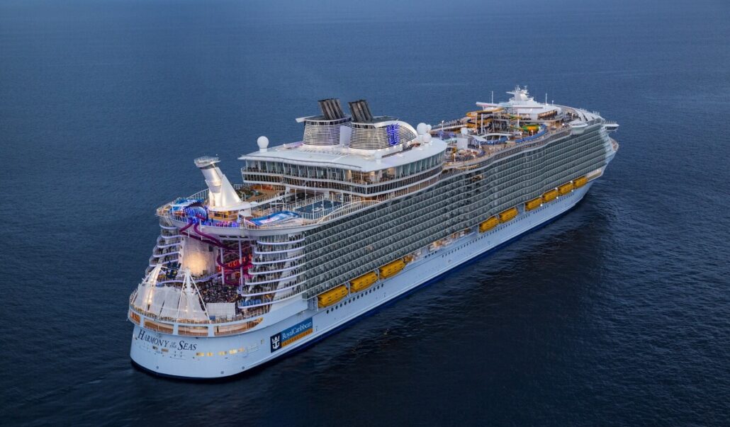 Aerial view of Royal Caribbean’s Harmony of the Seas sailing on calm blue water, with multiple decks, exciting ship upgrades like water slides, and various recreational areas visible on the upper levels.