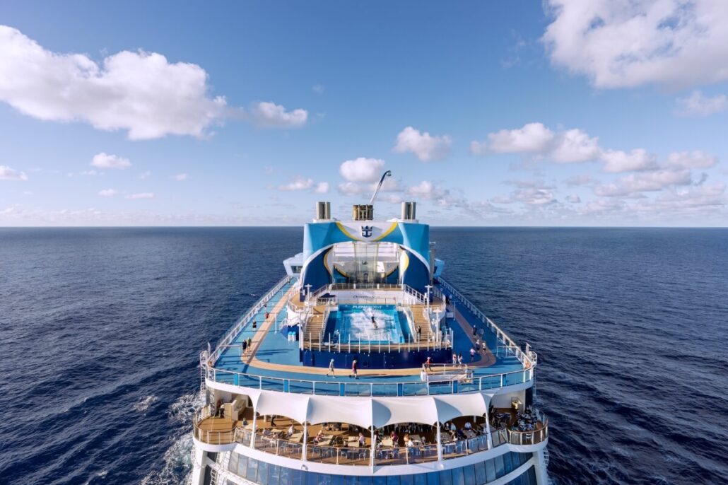 A Royal Caribbean cruise ship sails on the open sea under a blue sky with scattered clouds. The upper deck features upgraded amenities, including a swimming pool and leisure area, where passengers enjoy stunning ocean views.