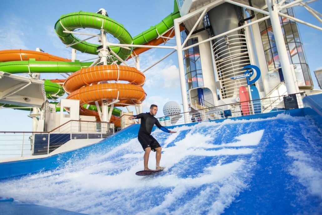 A man in a black wetsuit rides an artificial wave on a surf simulator. In the background, spiral green and orange water slides highlight Royal Caribbean’s exciting cruise ship upgrades.