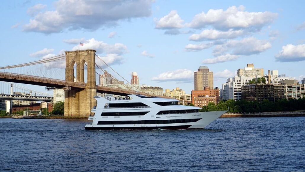 A modern white New York charter boat sails on a river with the Brooklyn Bridge and city buildings in the background under a partly cloudy sky.