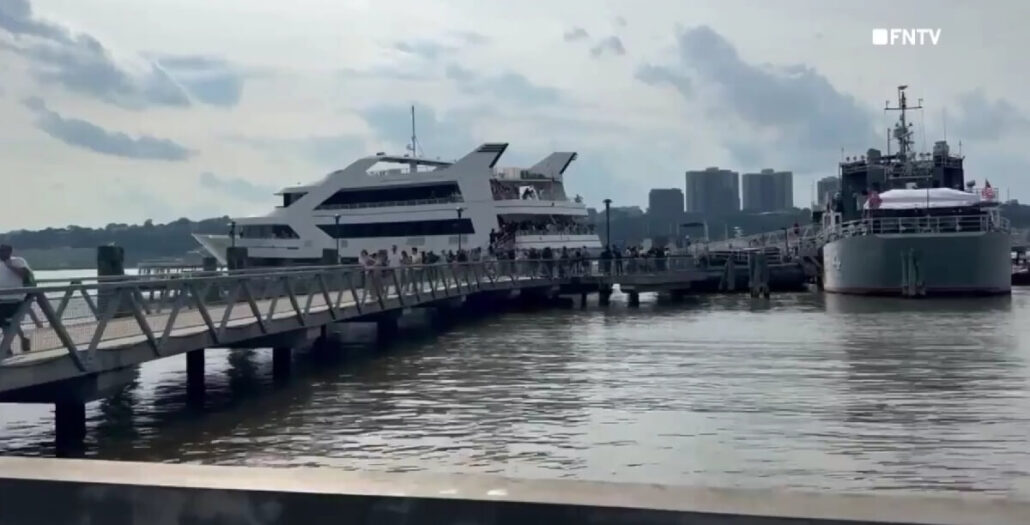 A pier in New York extends over the water as people walk toward two docked boats: a modern white charter boat and a gray ship. Buildings and a cloudy sky form the backdrop.