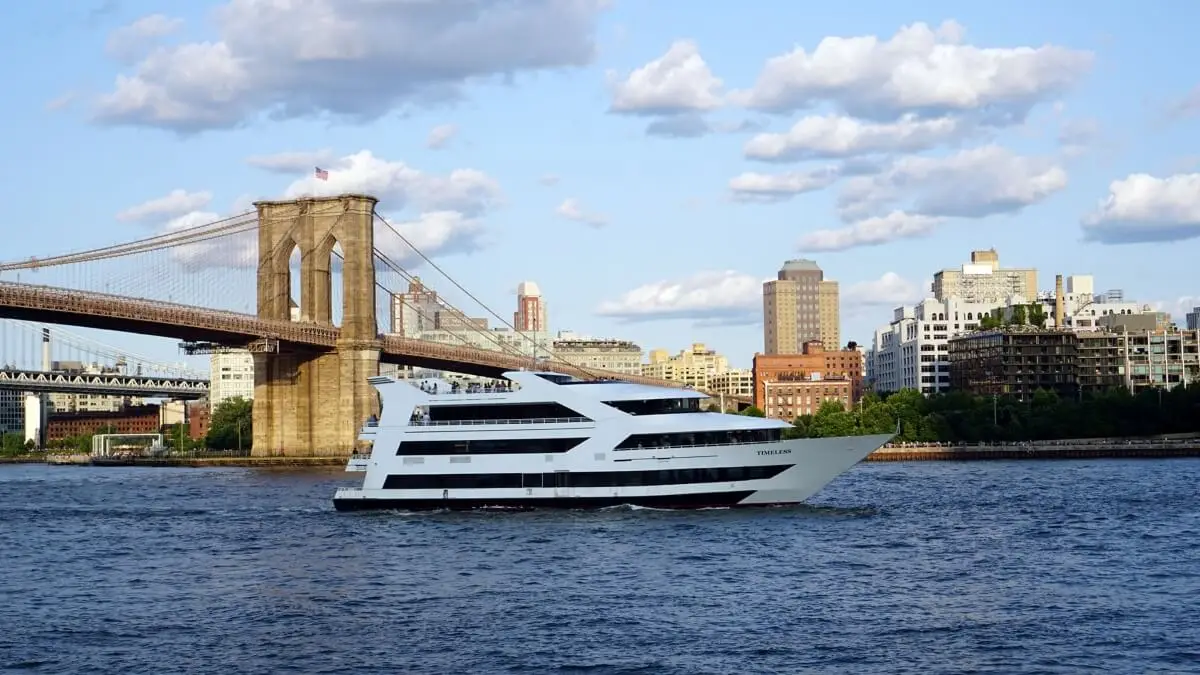A modern white New York charter boat sails on a river with the Brooklyn Bridge and city buildings in the background under a partly cloudy sky.