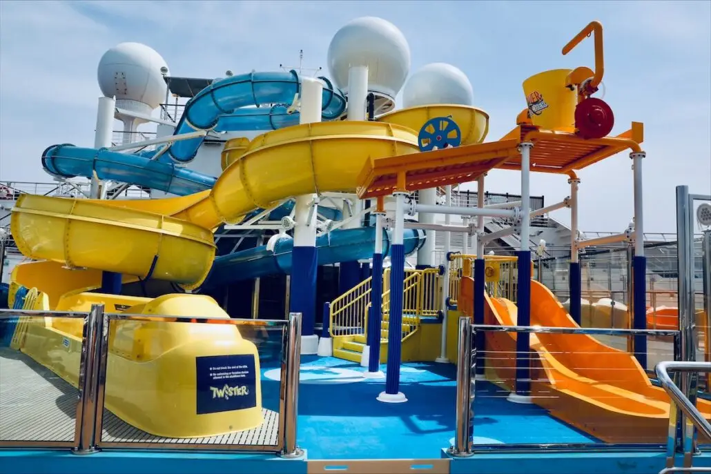 A colorful outdoor WaterWorks Park with yellow and orange slides, a blue spiral slide, and a large tipping bucket, surrounded by railings. This lively structure is located on the deck of Carnival Valor.