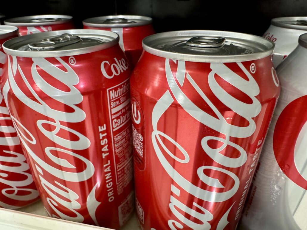 A close-up view of several red Coke cans with "Original Taste" text, stacked on a shelf, captures the fun spirit found on Carnival Ships—reminding you to grab your favorite drink for your cruise in September 2025. A Diet Coke can peeks in on the right.