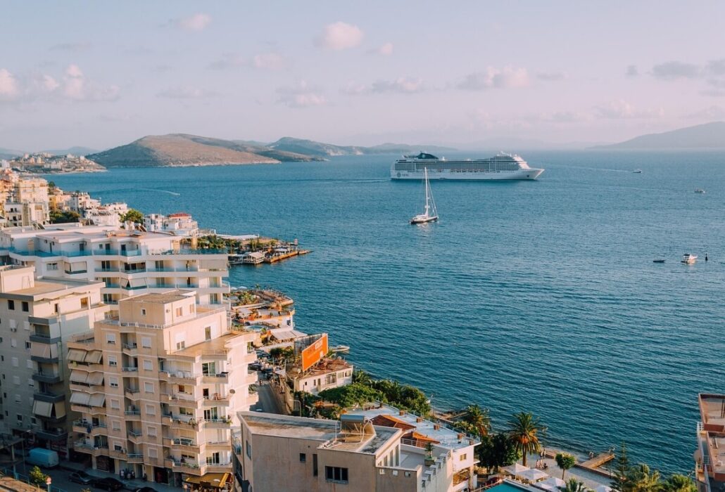 View of a coastal city with multi-story buildings in the foreground, a large cruise ship promising a memorable cruise experience and a sailboat in the blue water, with distant hills under a partly cloudy sky.