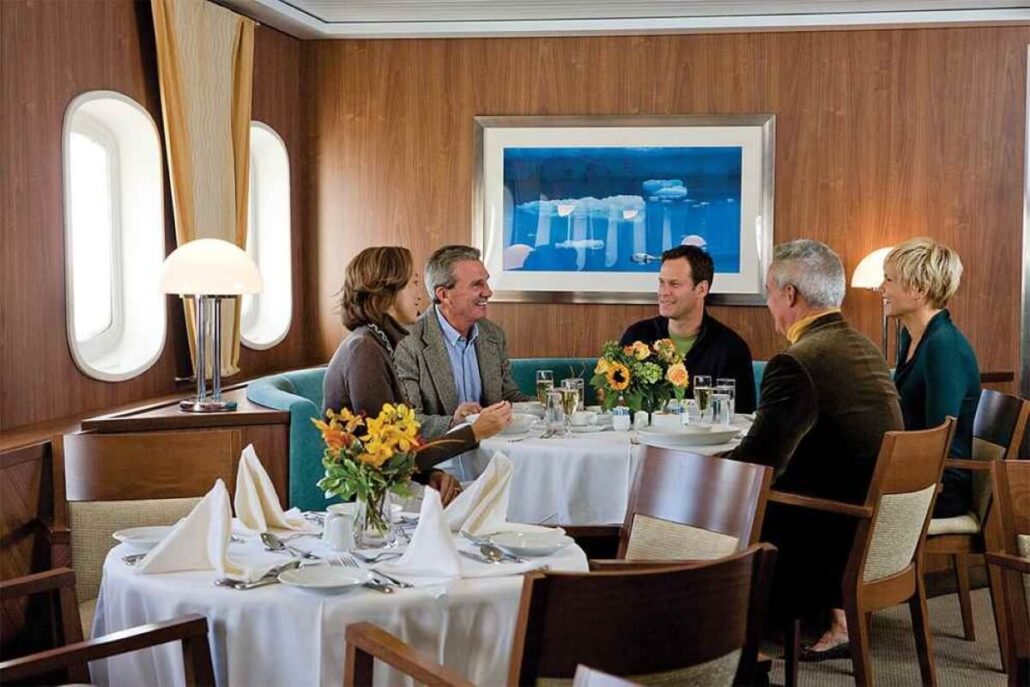 Five adults are sitting around a round table set for a meal in a dining room with wood-paneled walls, large windows, and framed artwork—reminiscent of an elegant Antarctica Expedition Cruise experience. The table has white linens and flower centerpieces.