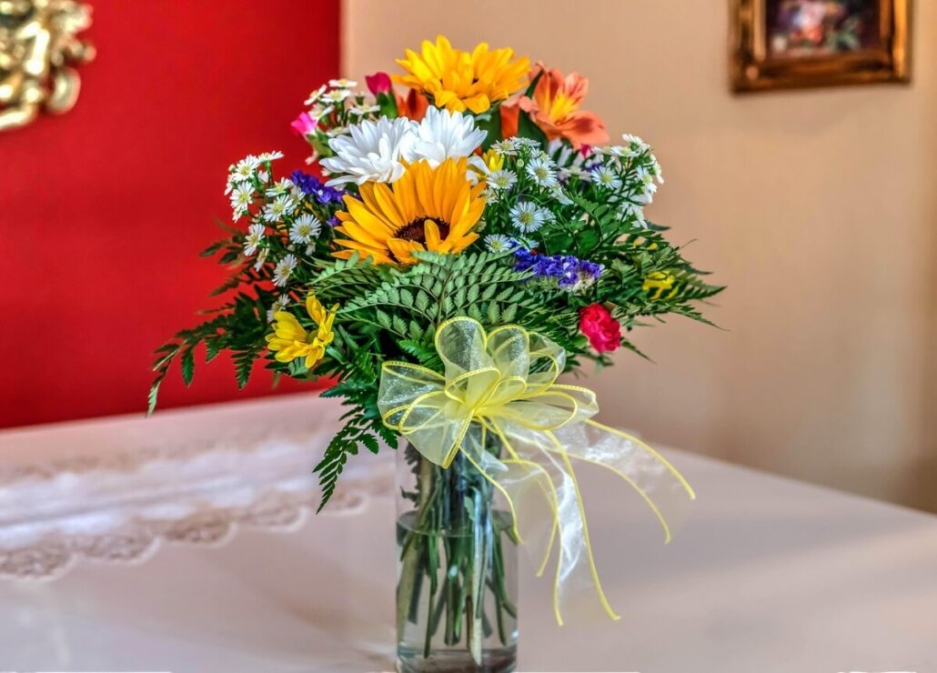 A clear glass vase holds a bouquet of assorted cruise flowers in yellow, white, and orange, with green foliage and a decorative yellow ribbon, placed on a white table against a red wall for ultimate cruise vibes.