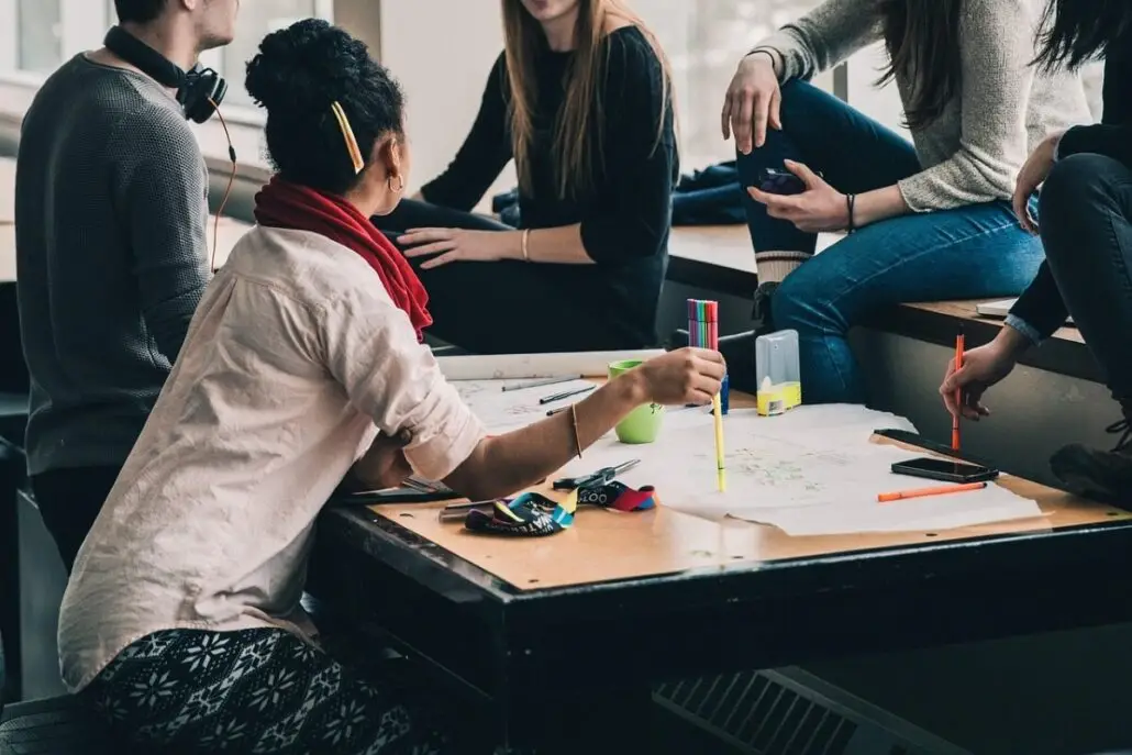 Five people sit and talk around a table covered with large sheets of paper, pens, and markers. They share tips on how to manage academics and discuss strategies for studying on the go while traveling. One person gestures with a marker as the group collaborates.