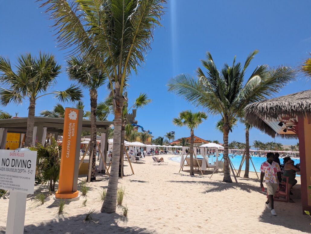 A sandy beach area at Celebration Key features palm trees, lounge chairs, and people relaxing near a large swimming pool under a clear blue sky. Signs highlight the Grand Opening, while shaded cabanas add comfort to the vibrant scene.
