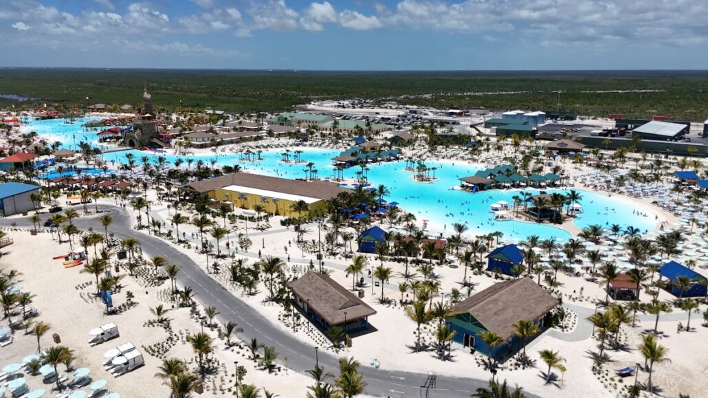 Aerial view of a large water park with an extensive artificial lagoon, palm trees, shaded cabanas, and pools—Capturing the excitement of a Grand Opening celebration amid buildings, greenery, and parking under a partly cloudy sky.