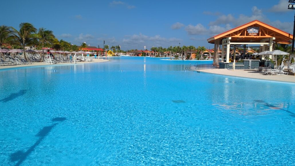 Large outdoor swimming pool with clear blue water, empty lounge chairs, palm trees, and a covered poolside bar under a wooden pavilion—get a behind the scenes look at this Celebration Key oasis on a sunny day with just a few clouds in the sky.