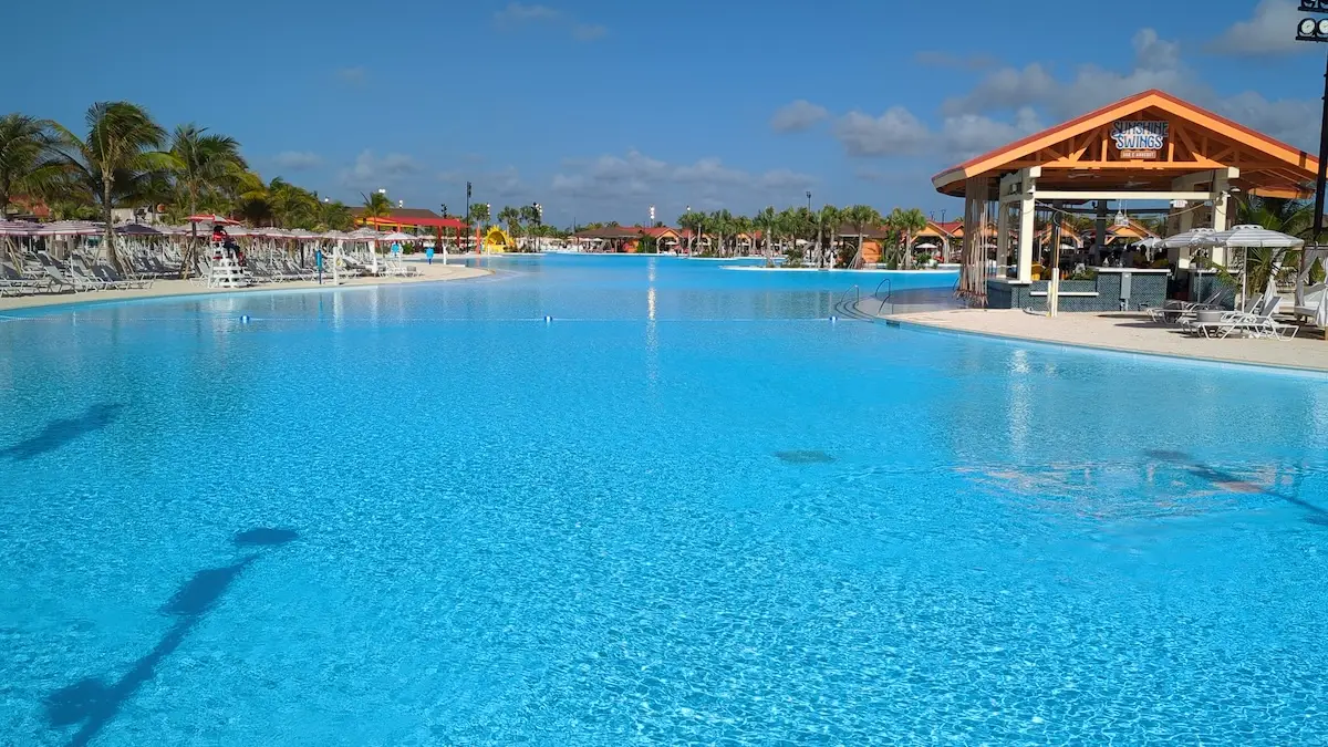 Large outdoor swimming pool with clear blue water, empty lounge chairs, palm trees, and a covered poolside bar under a wooden pavilion—get a behind the scenes look at this Celebration Key oasis on a sunny day with just a few clouds in the sky.