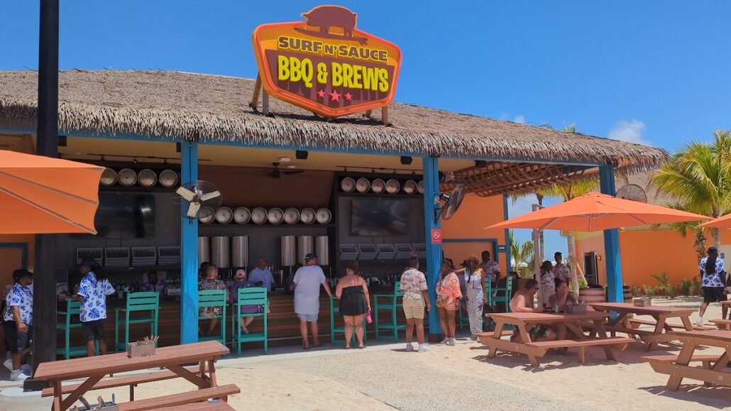 People stand in line at the Grand Opening of Surf & Sauce BBQ & Brews, an outdoor restaurant with a thatched roof. Orange umbrellas and picnic tables sit on the sandy ground nearby as guests enjoy a sunny day of delicious barbecue.