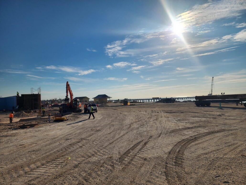 A construction site near a body of water under a bright, sunny sky. Workers, machinery, and vehicles are present, with buildings and a pier in the background. Tire marks are visible in the dirt—a true behind the scenes look at progress.