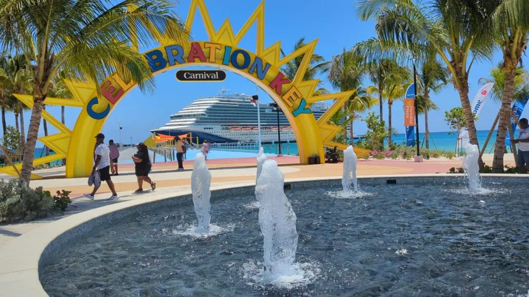 A cruise ship is docked near a beach behind a yellow and blue “Celebration Key” arch. Several people walk nearby, palm trees line the area, and water fountains are in the foreground during the Grand Opening festivities.
