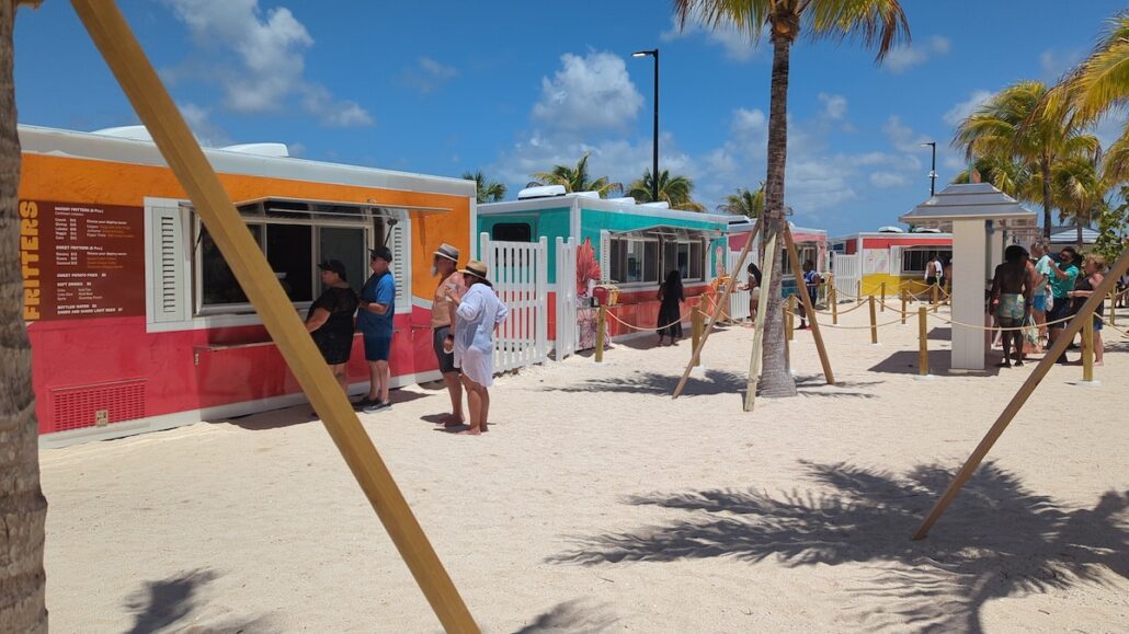 People stand in line at colorful food trucks on a sandy area with palm trees under a bright blue sky during the Grand Opening. Some people are ordering at the trucks while others wait, and a menu board is visible on one truck.