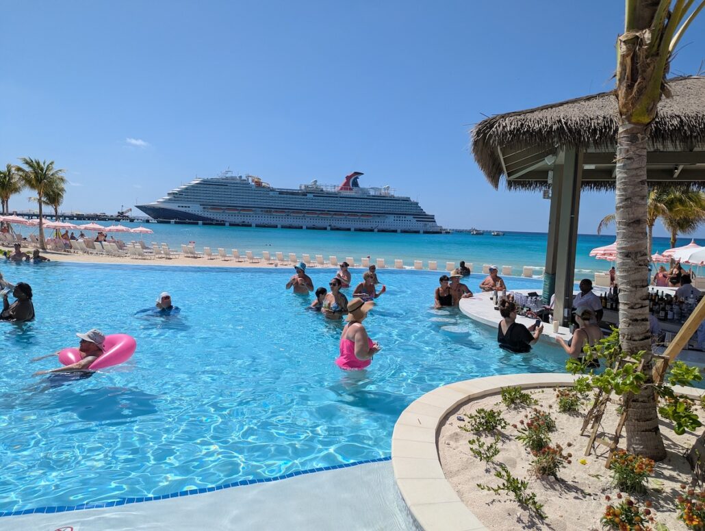 People relax and swim in a resort pool near Celebration Key beach, with a large cruise ship anchored in the background and clear blue skies overhead. Some guests enjoy drinks at a swim-up bar under a thatched roof during the grand opening festivities.