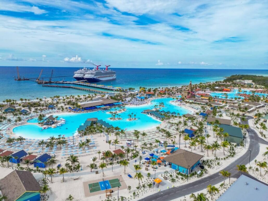 A large Carnival cruise ship is docked near a tropical beach resort with multiple pools, palm trees, and colorful buildings, ready to welcome 9,000 guests overlooking clear blue ocean water under a partly cloudy sky.