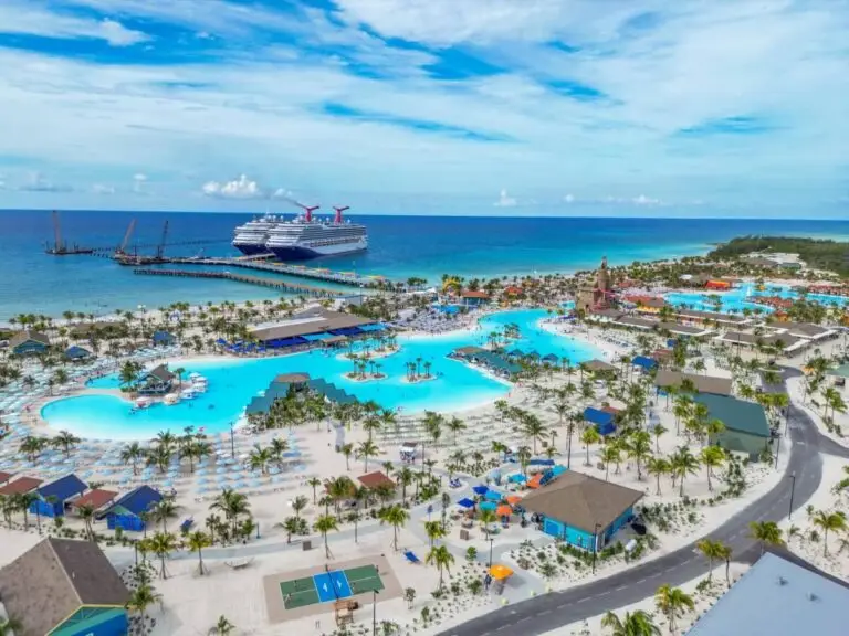 A large Carnival cruise ship is docked near a tropical beach resort with multiple pools, palm trees, and colorful buildings, ready to welcome 9,000 guests overlooking clear blue ocean water under a partly cloudy sky.